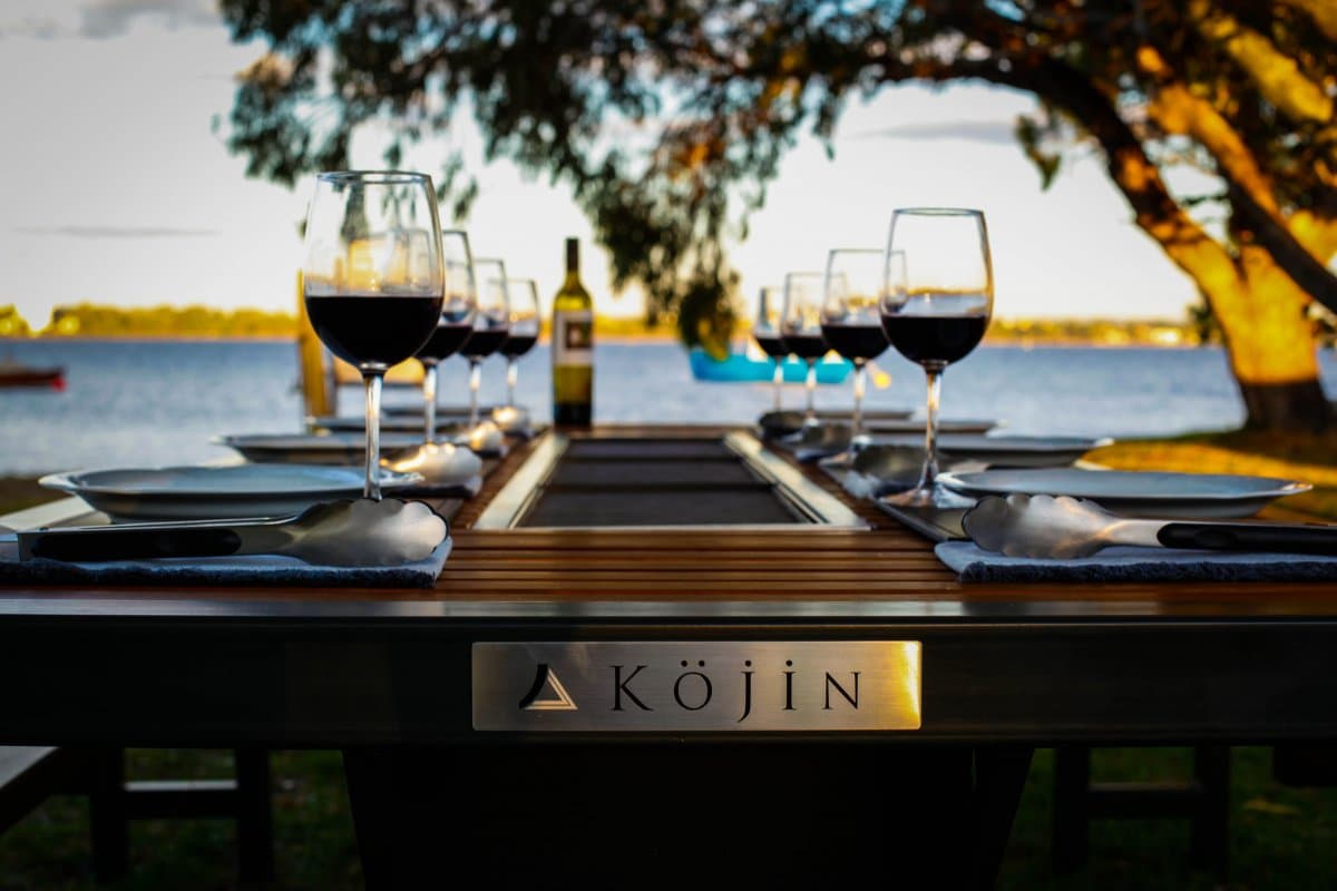 A Köjin alfresco dining table set for dinner beside the water at dusk, from the Jaime Alexander range