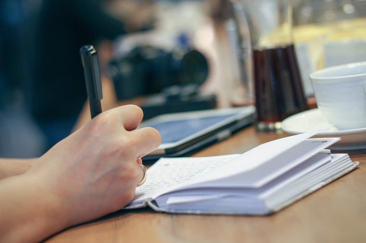 A hand writing in a notebook beside a coffee